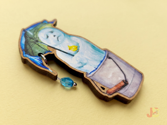 A 2.5 inch laser cut wooden brooch of a melting snowman holding and umbrella with a dangling blue glass bead raindrop and small yellow bird resting on his arm.