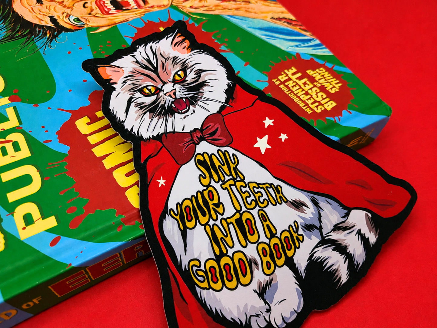 A close-up shot of a double-sided, die-cut bookmark featuring a Pallas Cat vampire wearing a red cape and bowtie.