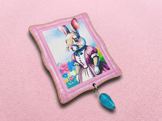 A 2.5 inch laser cut cherry wood brooch featuring an original illustration of a cottagecore style bunny in Victorian clothing. Attached is a blue glass bead.