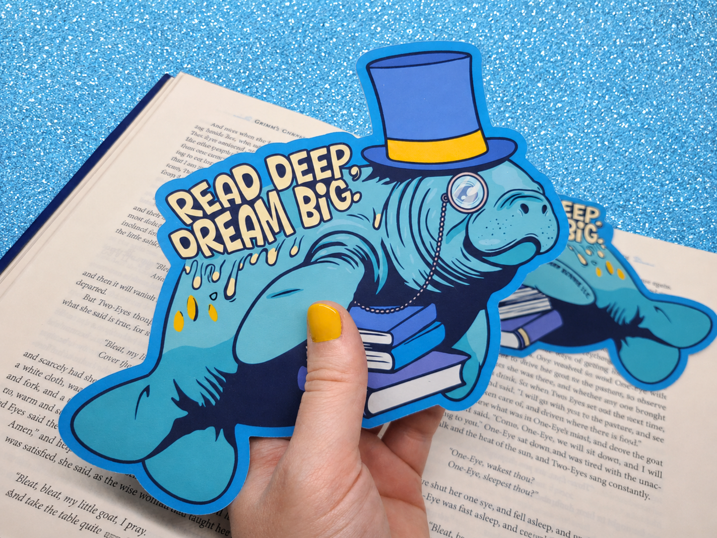 A die-cut bookmark being held up against an open book. It features an original illustration of a manatee wearing a top hat and monocle, carrying a stack of books. The bookmark includes the motivational quote "Read Deep, Dream Big."