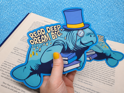 A die-cut bookmark being held up against an open book. It features an original illustration of a manatee wearing a top hat and monocle, carrying a stack of books. The bookmark includes the motivational quote "Read Deep, Dream Big."
