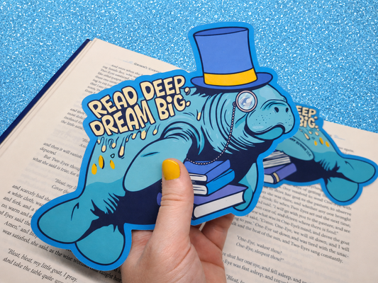 A die-cut bookmark being held up against an open book. It features an original illustration of a manatee wearing a top hat and monocle, carrying a stack of books. The bookmark includes the motivational quote "Read Deep, Dream Big."