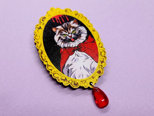 A 2.5 inch laser cut cherry wood brooch featuring an original illustration of a fluffy, grumpy Pallas cat with tiny vampire fangs. A red glass bead hangs from the bottom.