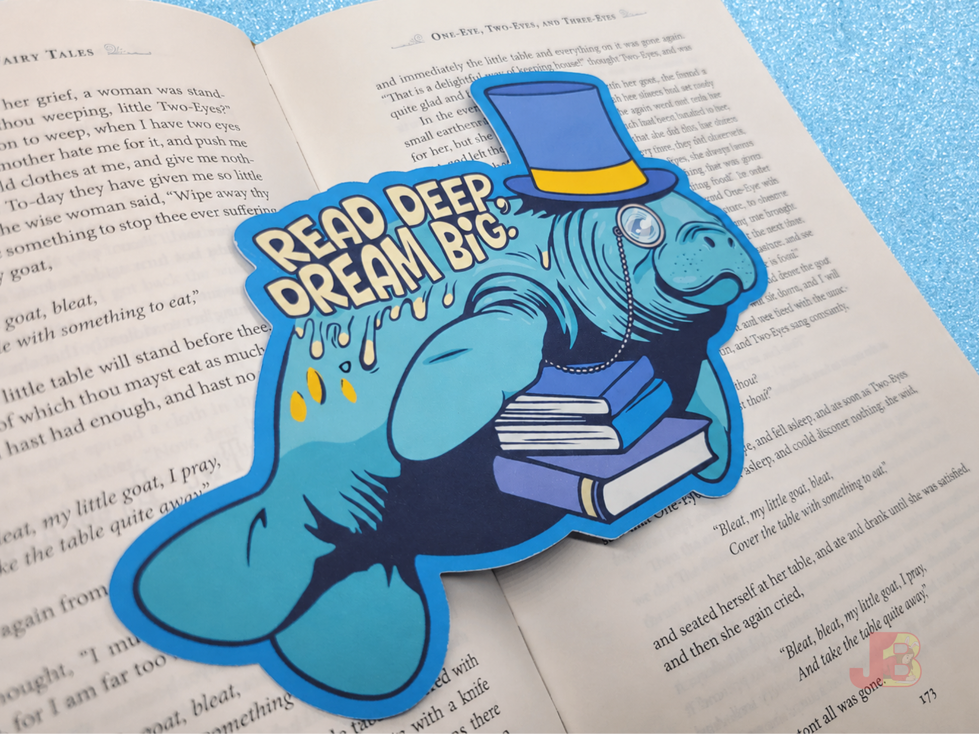 A die-cut bookmark resting on an open book. It features an original illustration of a manatee wearing a top hat and monocle, carrying a stack of books. The bookmark includes the motivational quote "Read Deep, Dream Big."