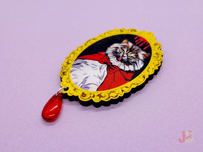 A 2.5 inch laser cut cherry wood brooch featuring an original illustration of a fluffy, grumpy Pallas cat with tiny vampire fangs. A red glass bead hangs from the bottom.