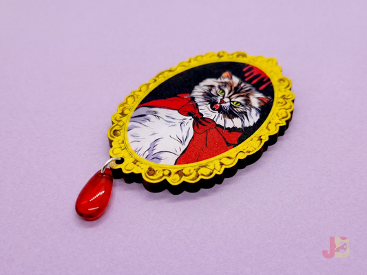 A 2.5 inch laser cut cherry wood brooch featuring an original illustration of a fluffy, grumpy Pallas cat with tiny vampire fangs. A red glass bead hangs from the bottom.