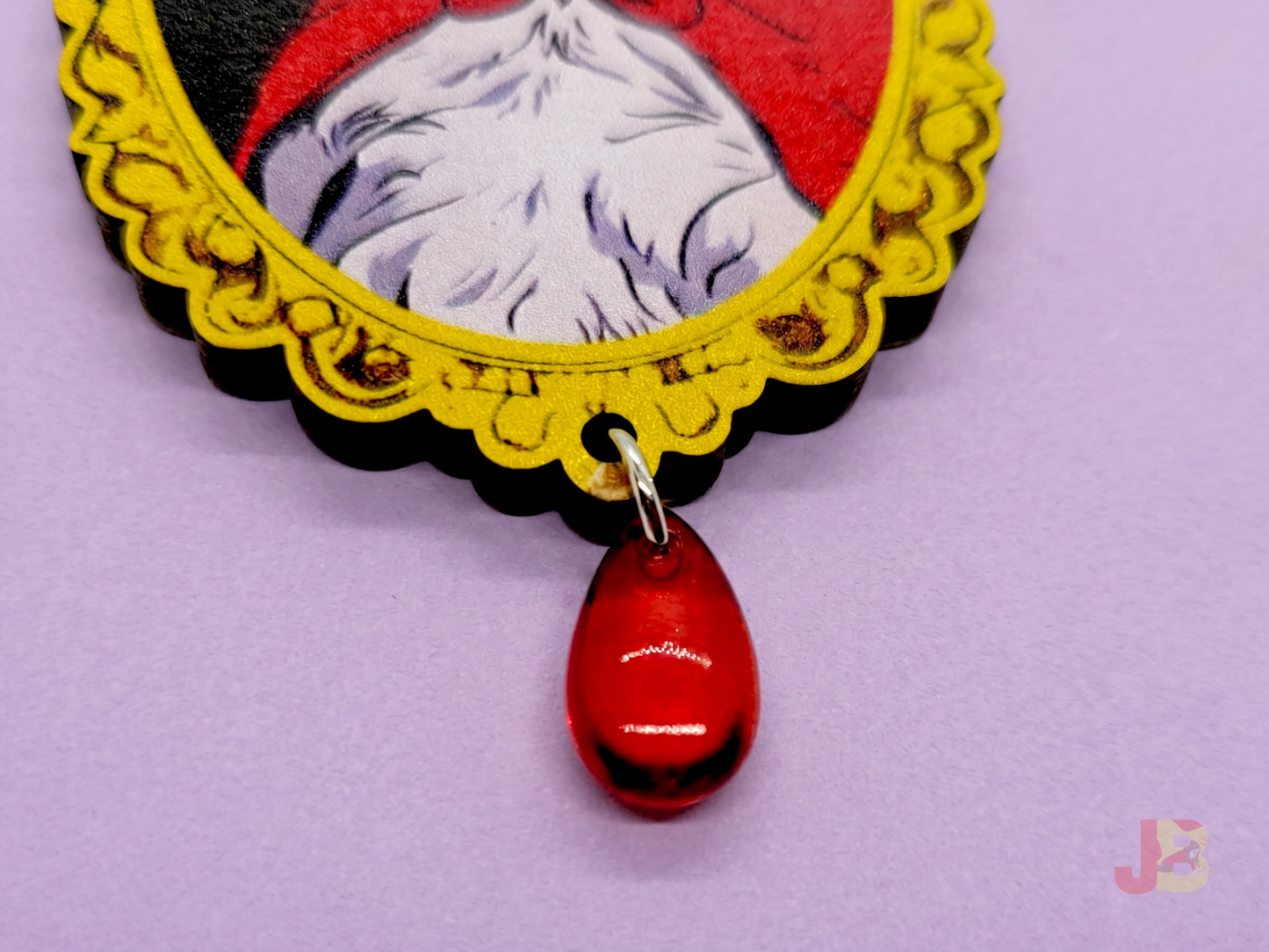 Up close of wooden brooch with a vampire Pallas cat illustration and red teardrop bead dangling from the bottom on a purple background.