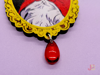 Up close of wooden brooch with a vampire Pallas cat illustration and red teardrop bead dangling from the bottom on a purple background.