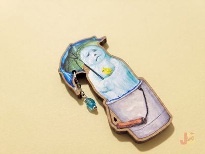 A 2.5 inch laser cut wooden brooch of a melting snowman holding and umbrella with a dangling blue glass bead raindrop and small yellow bird resting on his arm.