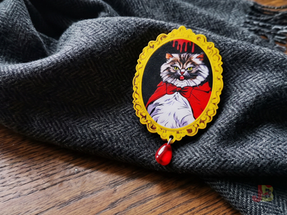 A 2.5 inch laser cut cherry wood brooch featuring an original illustration of a fluffy, grumpy Pallas cat with tiny vampire fangs. A red glass bead hangs from the bottom.