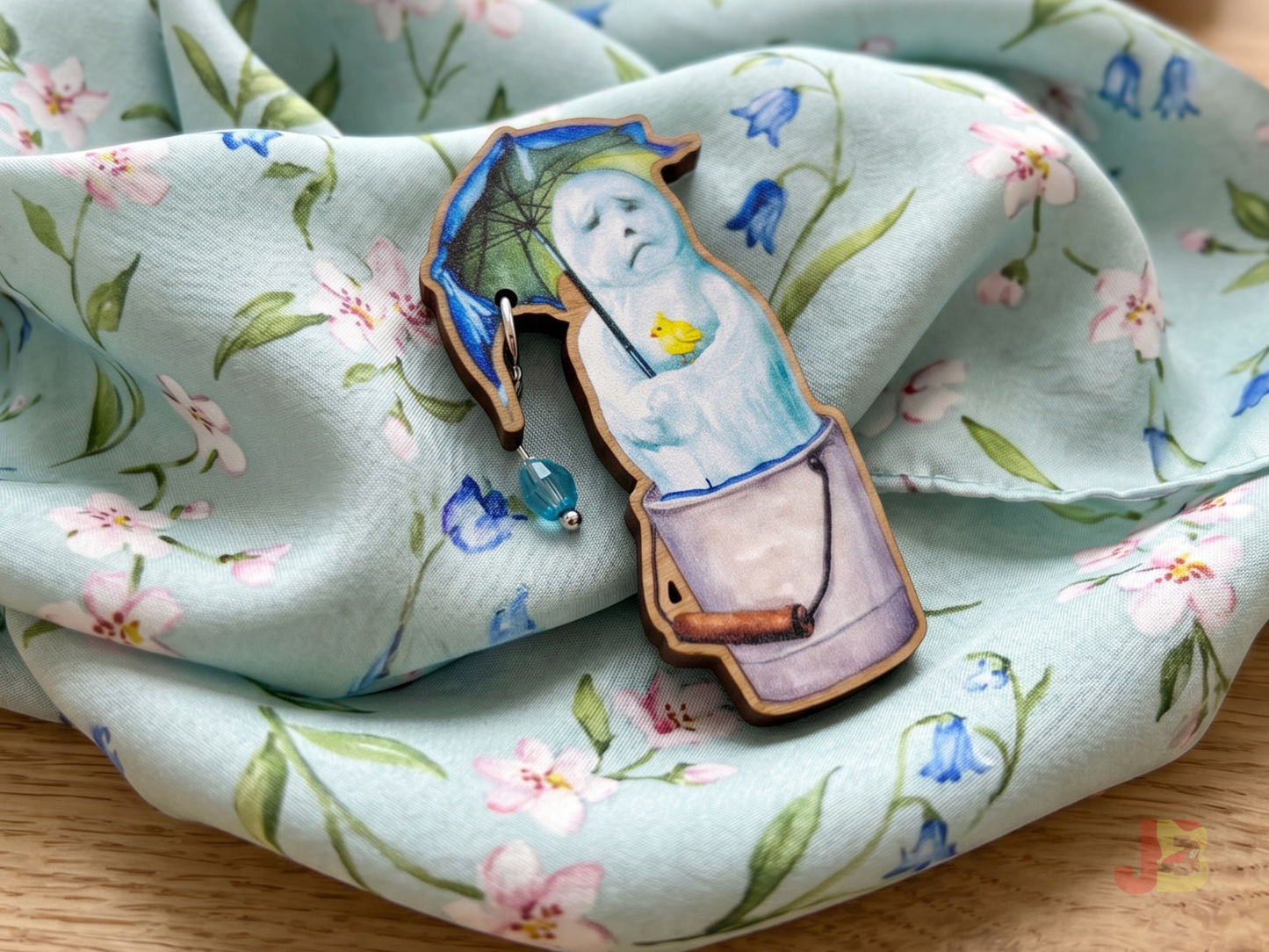 The melting snowman wooden brooch resting on a spring themed scarf laying on a table.